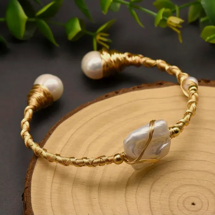 Open rigid cuff of gold-tone beads with a baroque pearl at each end, on wood.